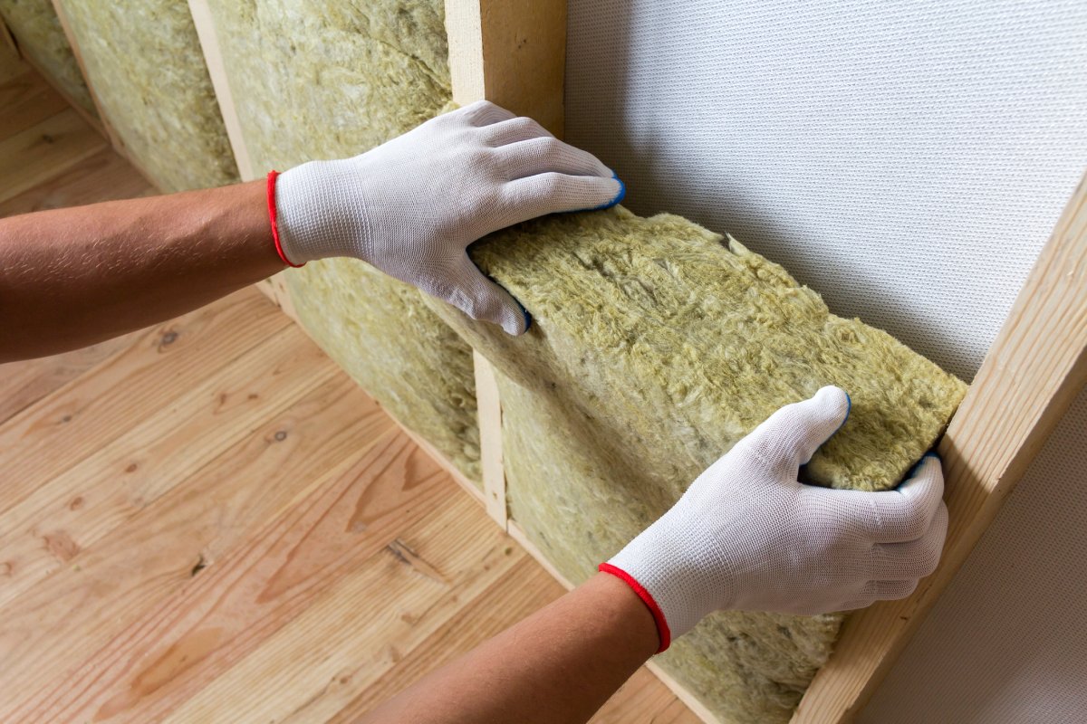 Hands wearing white gloves install yellow mineral wool insulation in a wooden frame wall. The setting shows a smooth wooden floor, conveying a sense of careful construction.